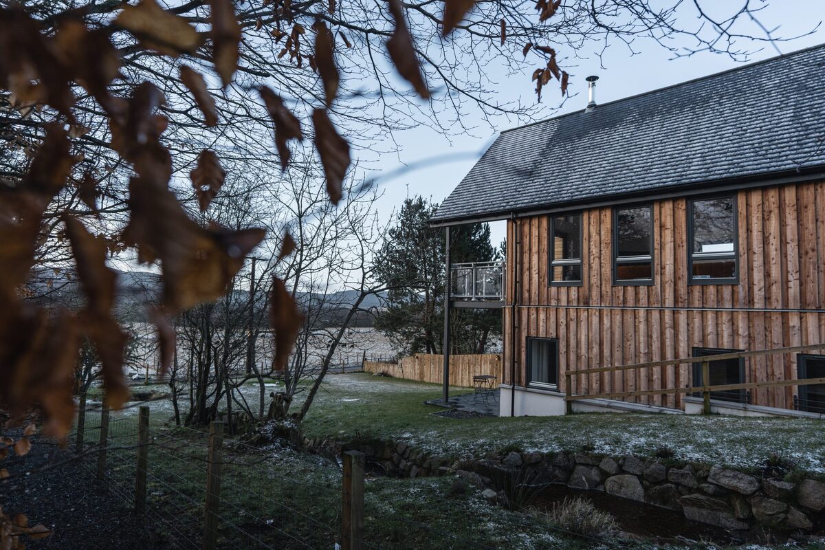 Timber-clad lodge with balcony overlooking a loch, framed by bare branches