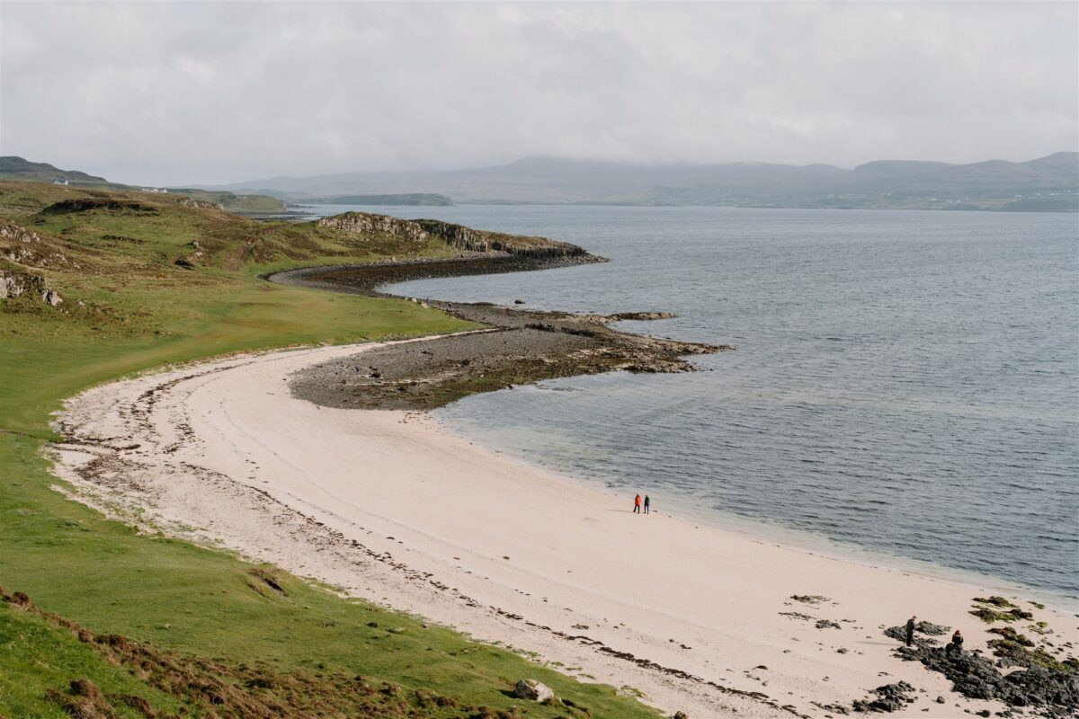 Two walkers cross a curved white sand beach beside green coastal hills