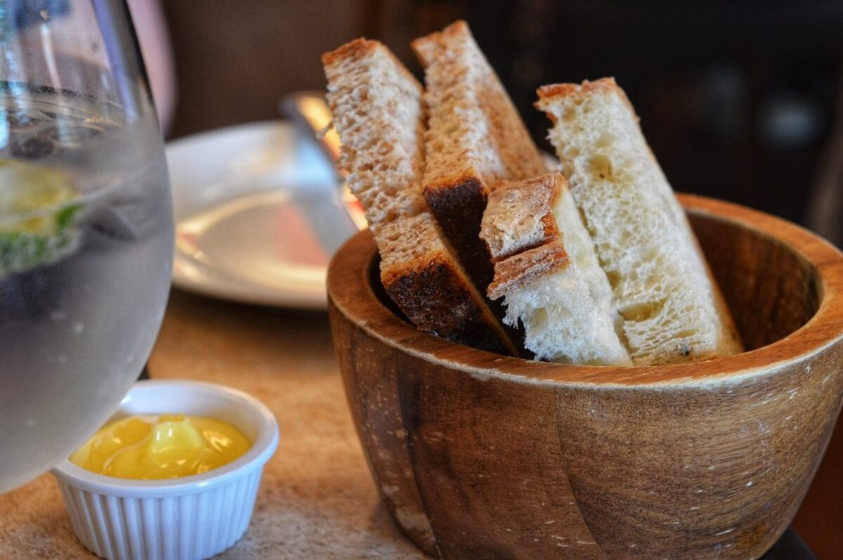 Sliced bread in a wooden bowl beside butter and a gin glass