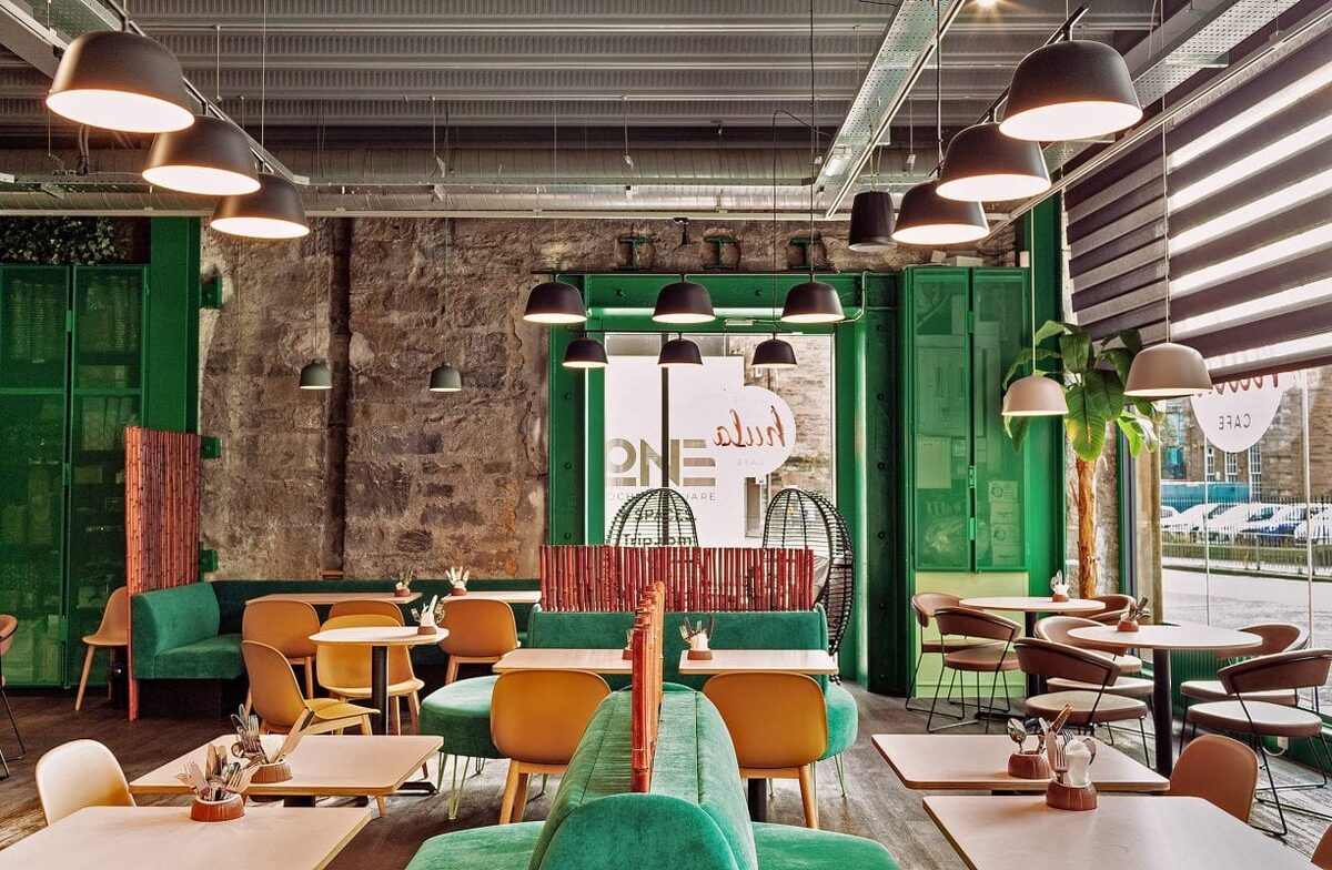 Green booths and mustard chairs fill a café with exposed stone walls and black pendant lights