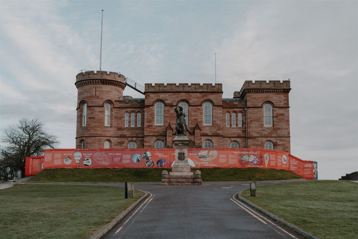 Inverness Castle's red sandstone towers rise behind a statue and hoarding-lined path