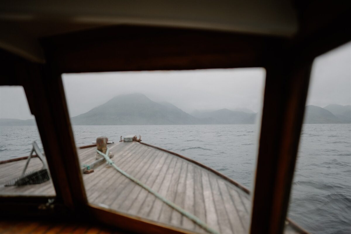 Wooden boat deck framed by cabin window, misty hills across the loch