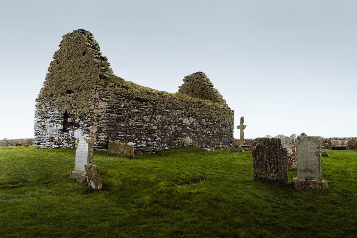 Ruined stone chapel with gabled walls surrounded by weathered gravestones and a Celtic cross