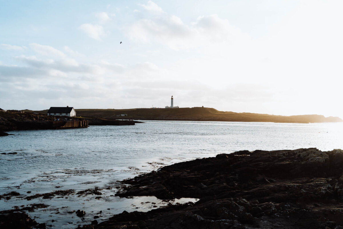 The lighthouse on Orsay small on the skyline, dark rocks and still water in front.