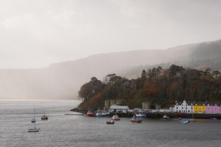 Colourful houses line Portree harbour beneath misty, tree-covered hillside, with boats moored offshore