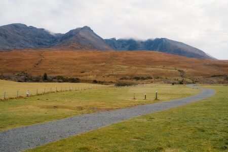 Gravel path curves through green fields towards mist-covered mountain peaks