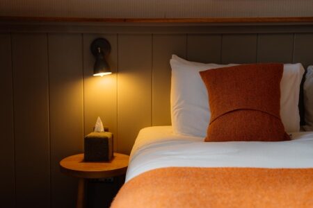 Bedside lamp lit above a round table with tissue box, orange cushion on white pillows