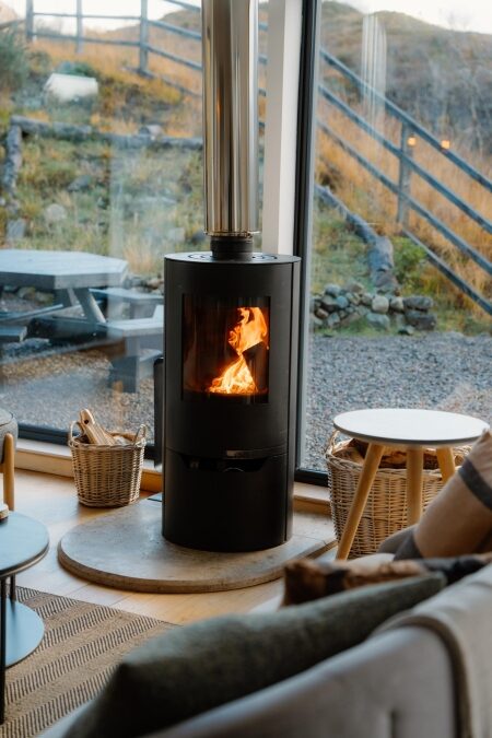 Wood-burning stove alight beside floor-to-ceiling windows overlooking a hillside path