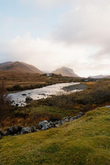 A river winds through moorland with a white cottage below misty hills
