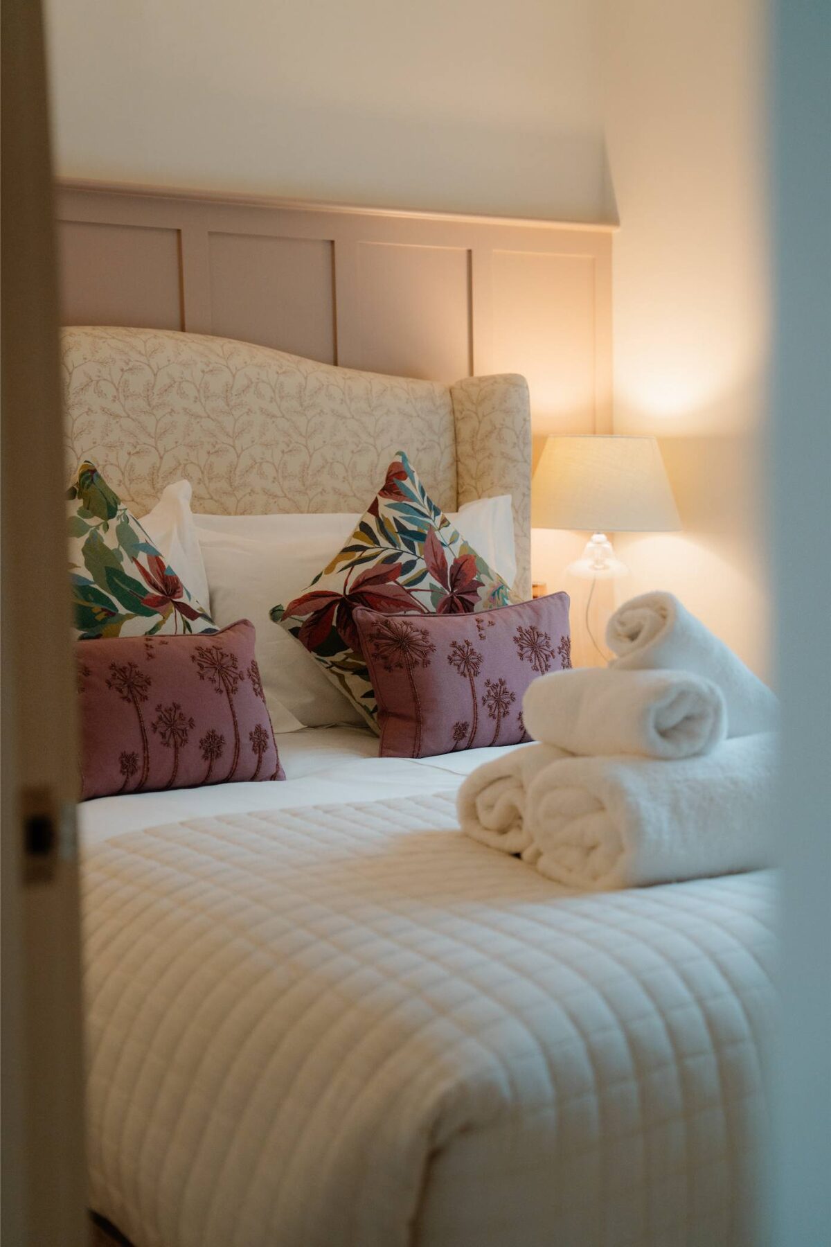 A bed with a cream tufted headboard and floral and burgundy cushions holds folded towels, seen through an open doorway.