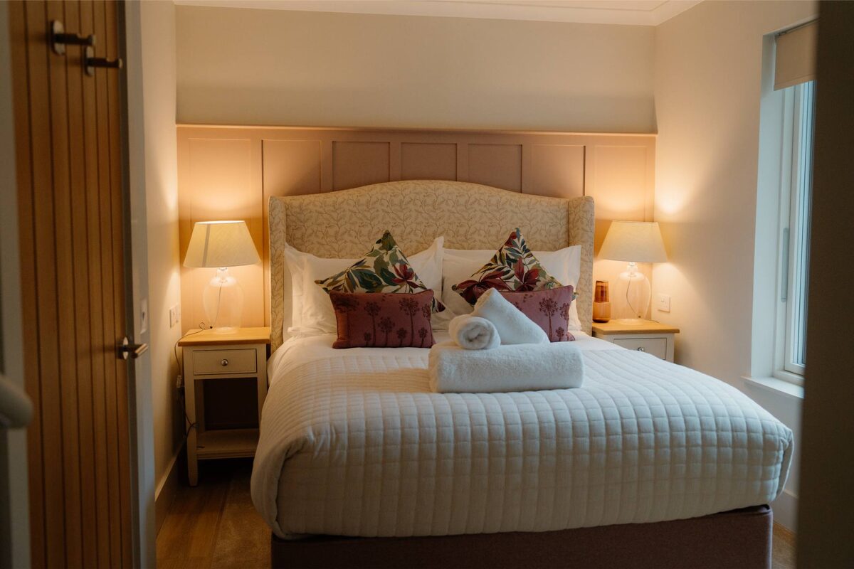A bed with a cream upholstered headboard and burgundy and floral cushions sits between twin bedside lamps, a rolled towel resting on the linen.