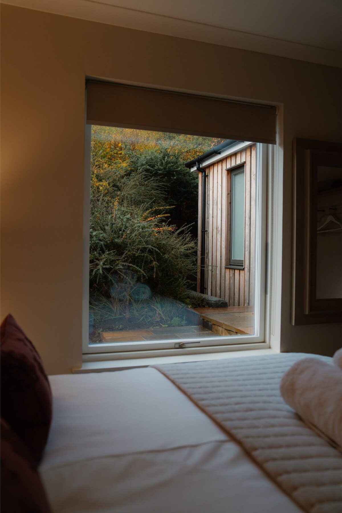 A bedroom window looks out to a timber-clad outbuilding and shrubs at dusk, the edge of a bed in the foreground.