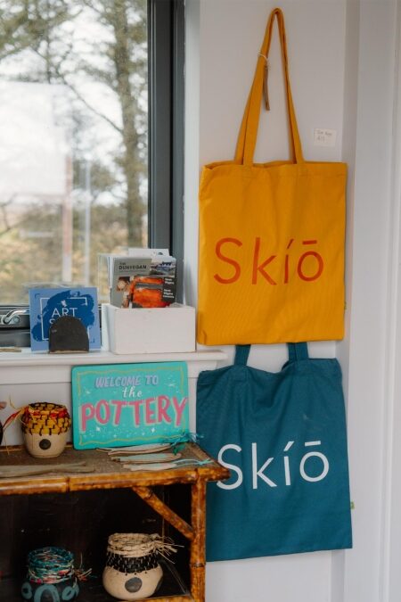 Yellow and teal Skío tote bags hang beside a "Welcome to the Pottery" sign