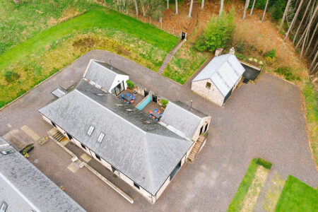 An aerial view shows a long stone steading with a slated roof and a rooftop hot tub, a smaller outbuilding and gravel courtyard beside it.