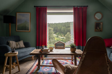 Sofas flank a window framed by red curtains looking out to a garden and outbuilding, a coffee table set with candles and a small house-shaped ornament between them.
