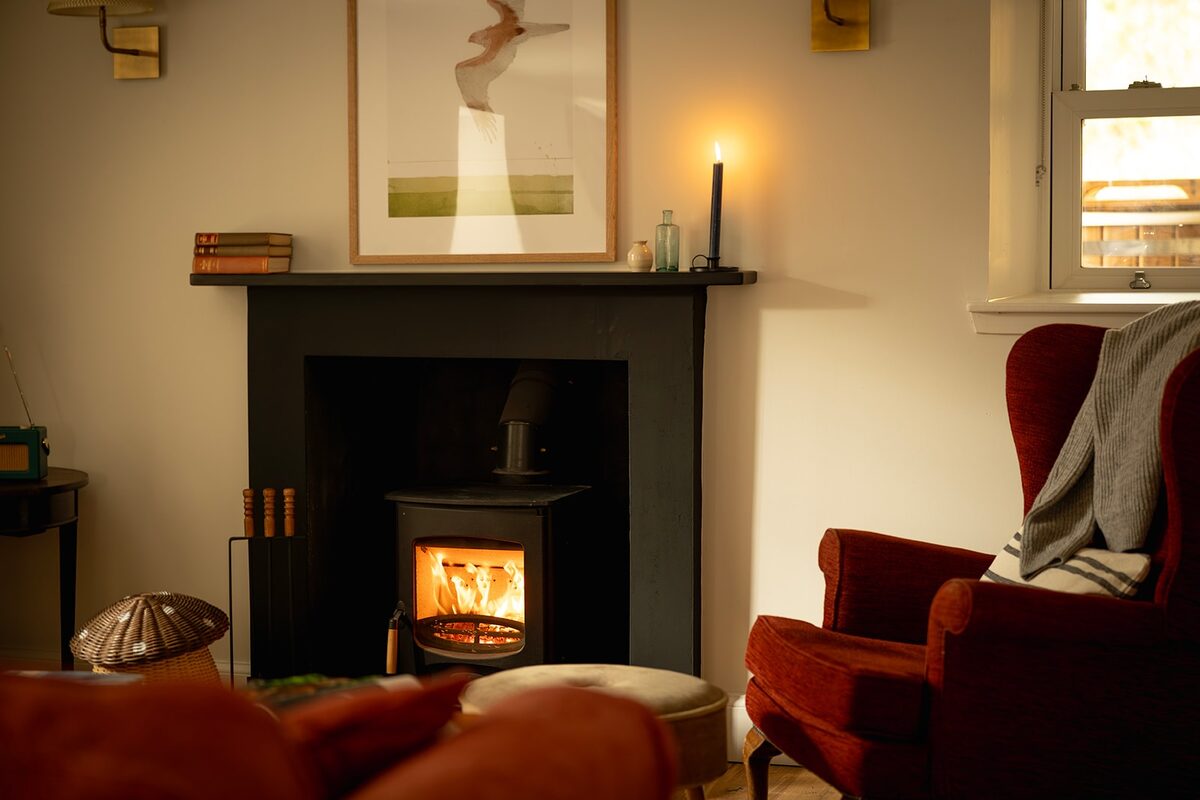 A wood burning stove glows in a black fireplace below a framed print, a lit candle and stacked books on the mantel beside it.