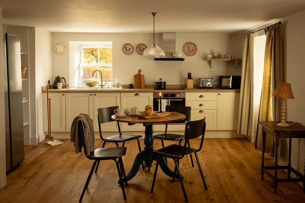 A round table and mismatched chairs stand before a kitchen with cream units, decorative plates hung on the wall above the window.