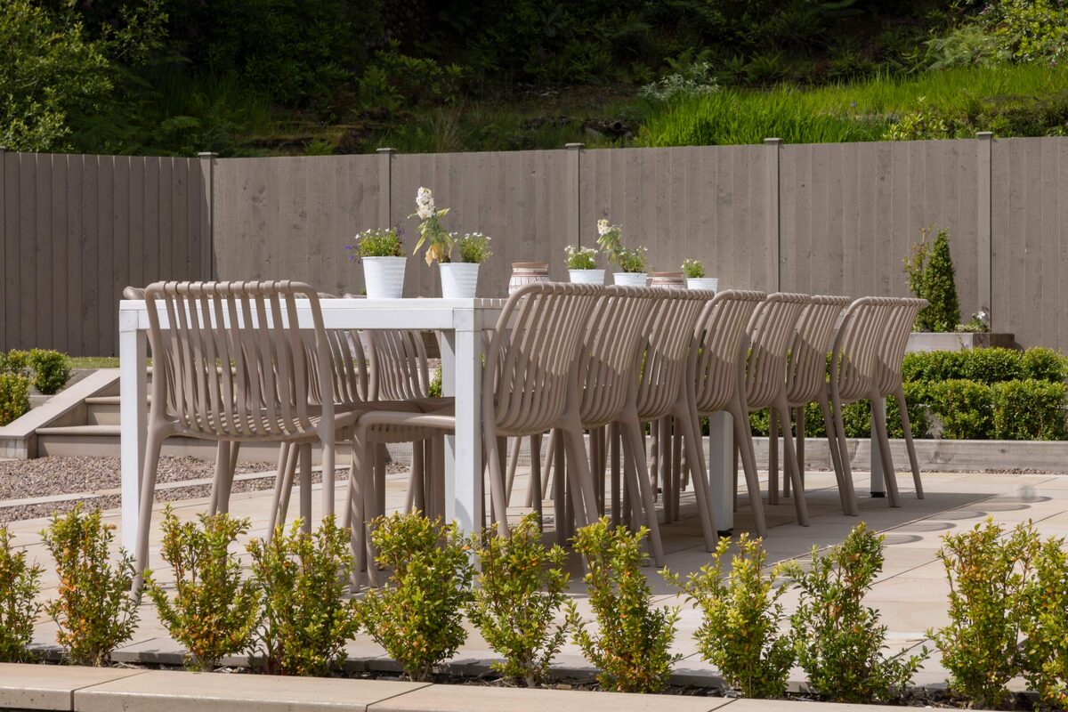 A long white outdoor table is set with potted plants and surrounded by pale stacking chairs on a gravel patio, a fence and hedge behind.