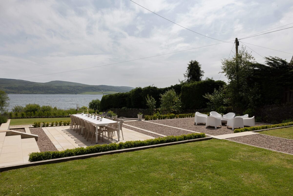 An elevated view shows a gravel patio with a dining table and white outdoor seating beside a lawn, a loch and hills stretching into the distance.
