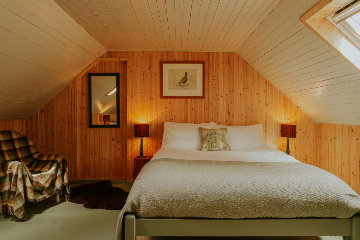 A bed with white linen sits beneath a sloped ceiling, a plaid armchair to the left and two red lamps lit on the bedside tables either side.