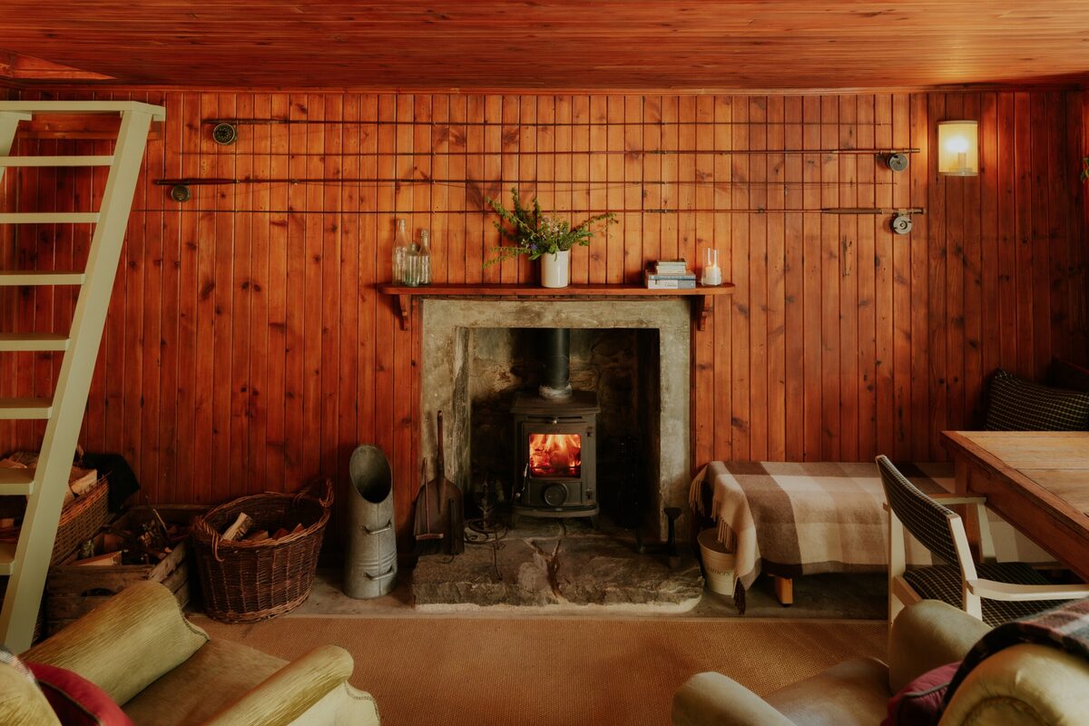 A wood burning stove burns in a stone fireplace beneath a mantel with a potted plant, timber-panelled walls and a log basket to the side.