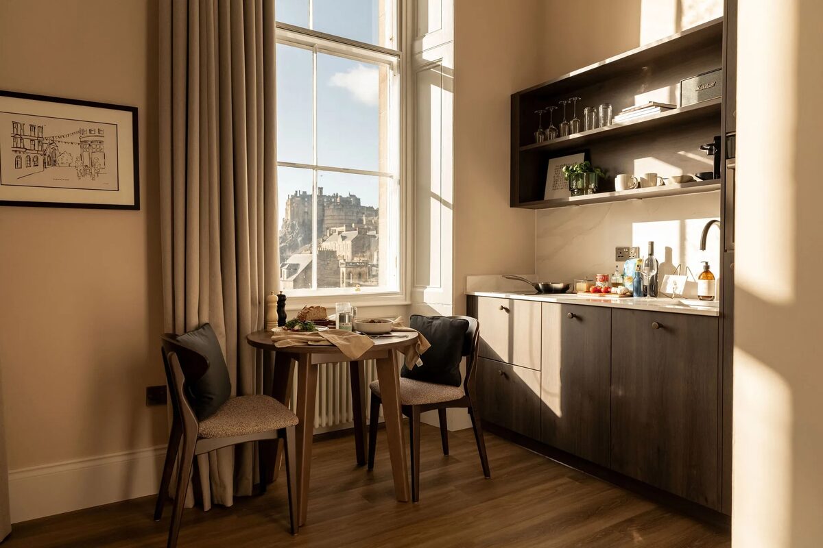 A round table set with breakfast items stands before a tall window with a castle view, a kitchenette with open shelving to the right.