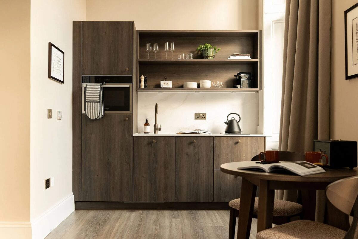 A kitchenette with dark wood cabinetry and open shelving of glassware stands beside a round table with an open book, curtains framing the window.