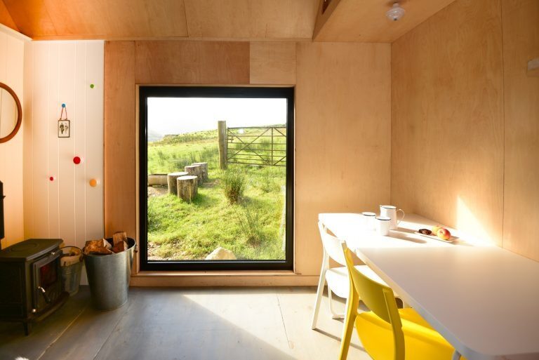 A yellow chair sits at a white table set with tea and fruit, a large window framing a fenced field beyond a wood burning stove.