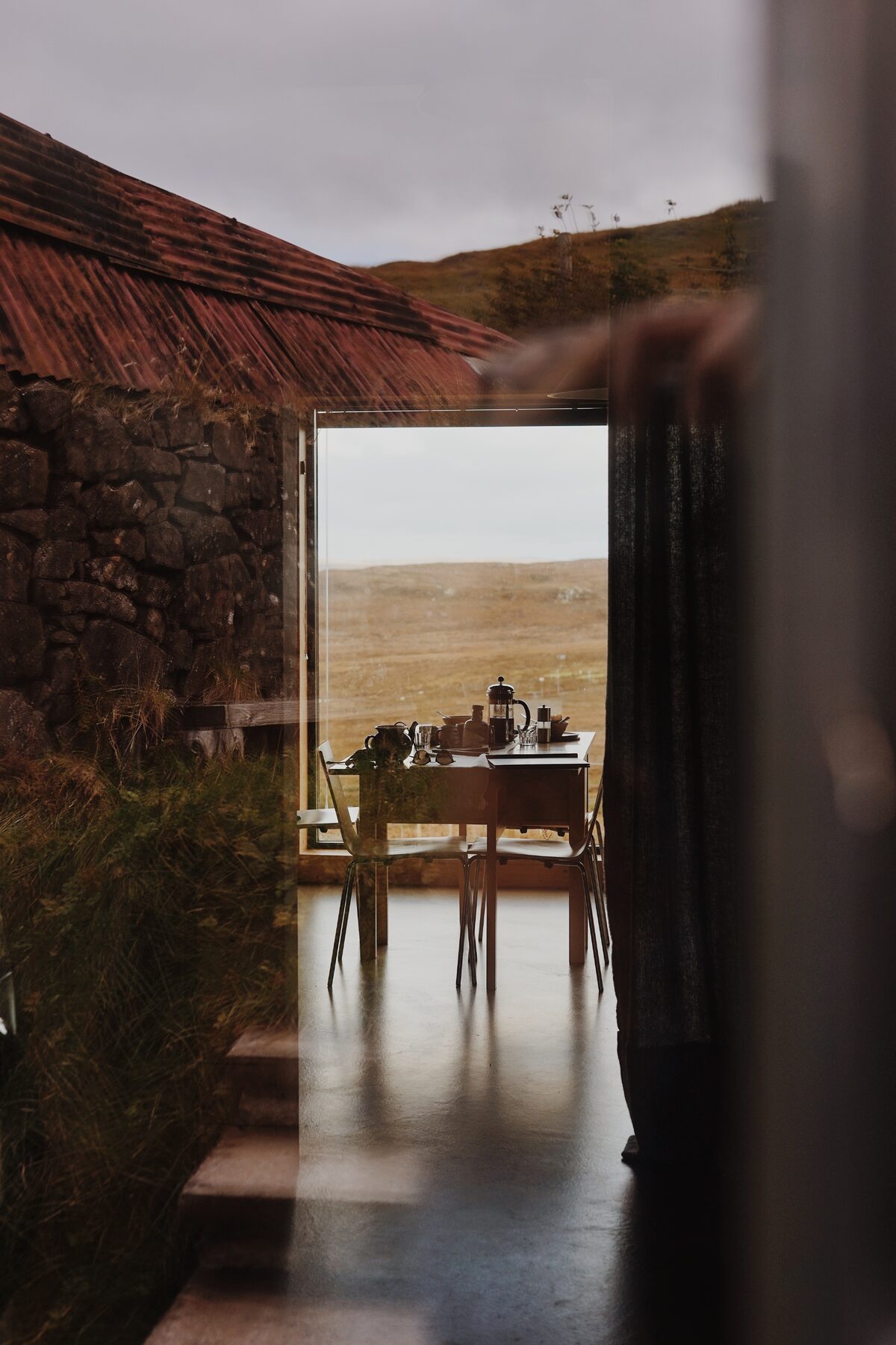 A stone building with a rust-coloured corrugated roof frames a glazed dining area, a table set with a coffee pot and mugs looking onto open moorland.