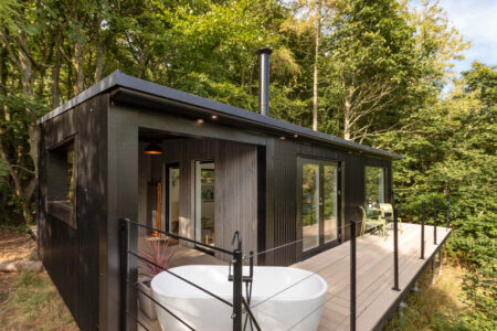 The black timber cabin's exterior, with a chimney above the roofline and a freestanding bath standing on the decking below.