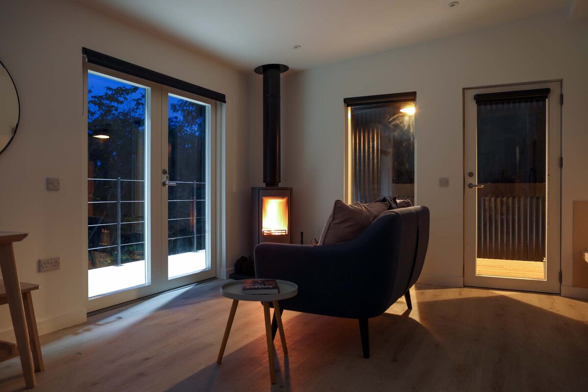 A wood burning stove lit at dusk beside a dark armchair, glazed doors standing open onto the deck.