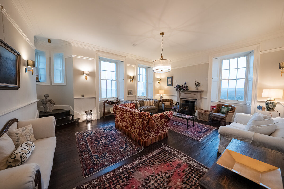 A patterned sofa and armchairs surround a low table in a bay-windowed sitting room, a fireplace and further seating visible beyond.