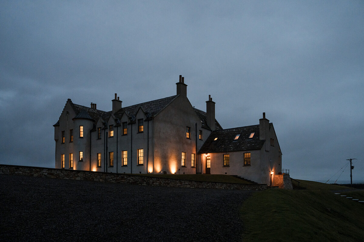 A white mansion with tall chimneys glows with lit windows at dusk, set on a grassy rise above a gravel drive.