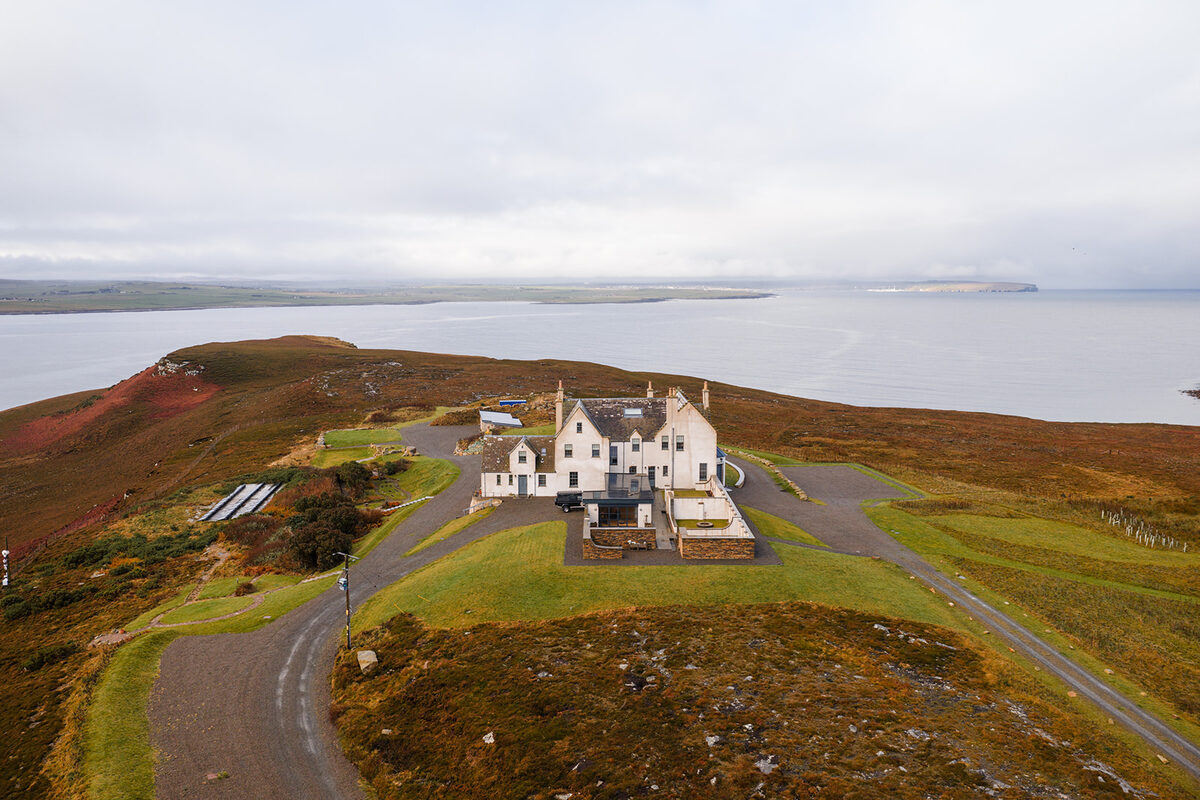 An aerial view shows a white mansion on a grassy headland above a wide sea loch, a road winding through heather-covered moorland.