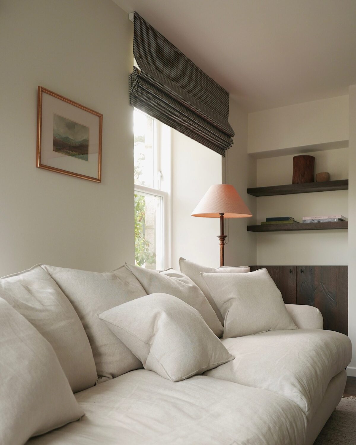 A grey linen sofa piled with cushions sits before a curtained window, an orange lamp and floating shelves with ornaments in the alcove behind.