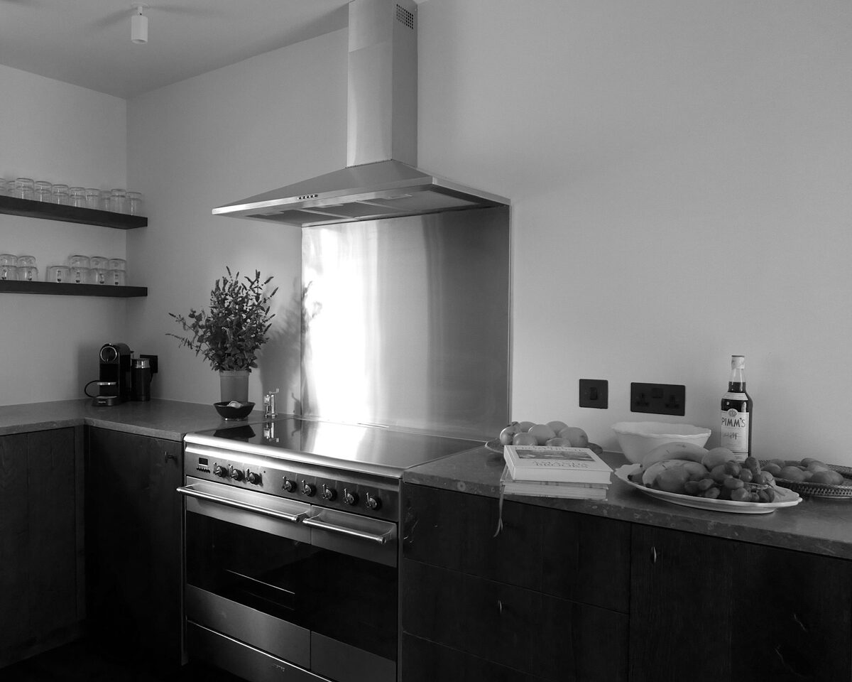 A range cooker with a stainless steel splashback and hood stands in a dark kitchen, plates of scones and pastries laid on the counter beside a bottle of wine and a jug of flowers.