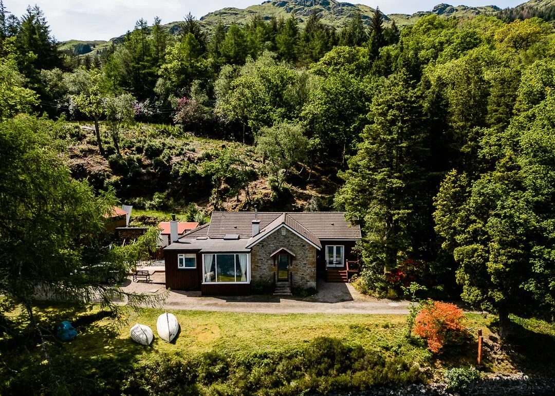 A stone cottage with a slated roof sits nestled among dense woodland, two paddleboards resting on the lawn in front.