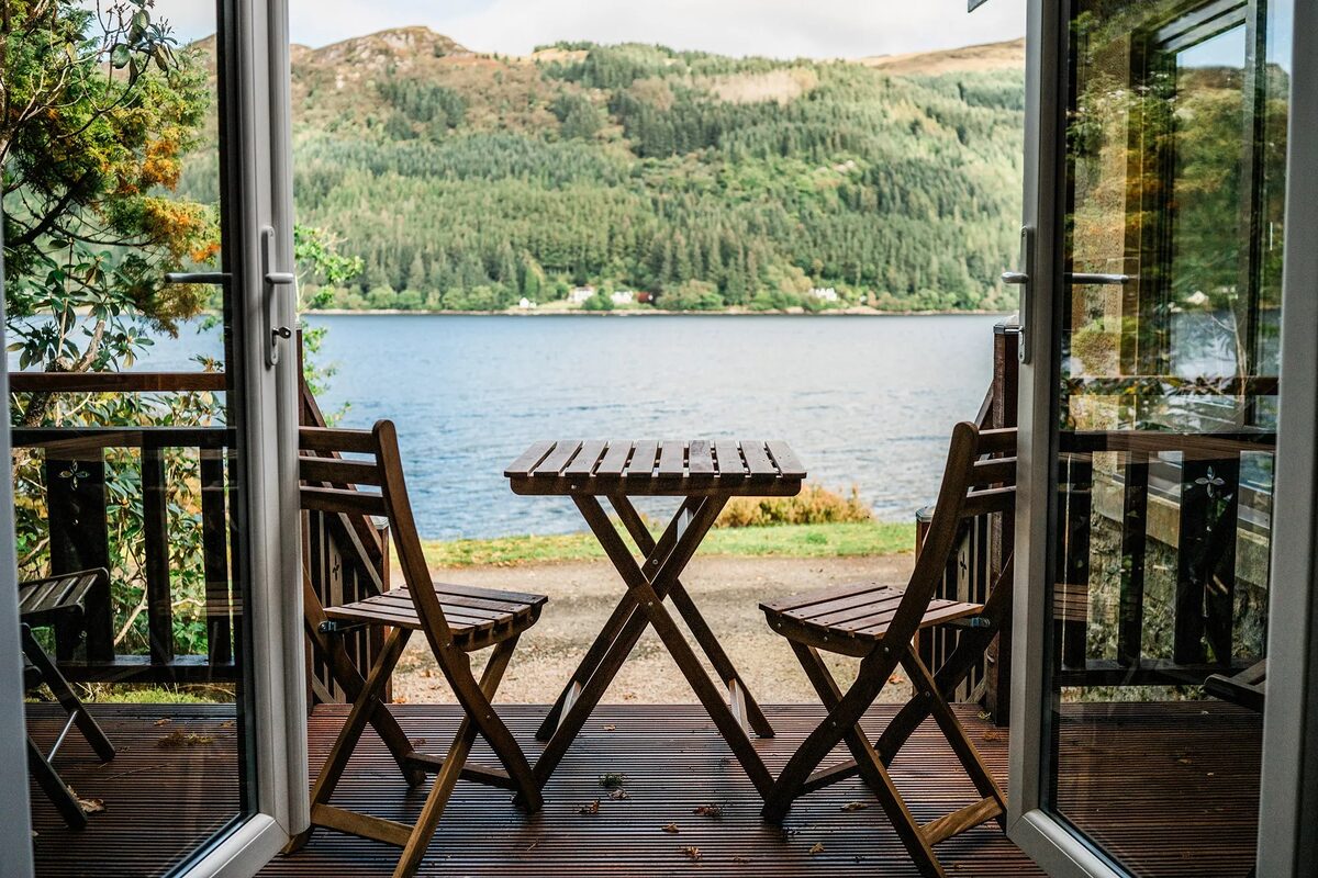 Two folding wooden chairs and a table stand on a deck looking out over a loch towards wooded hills.