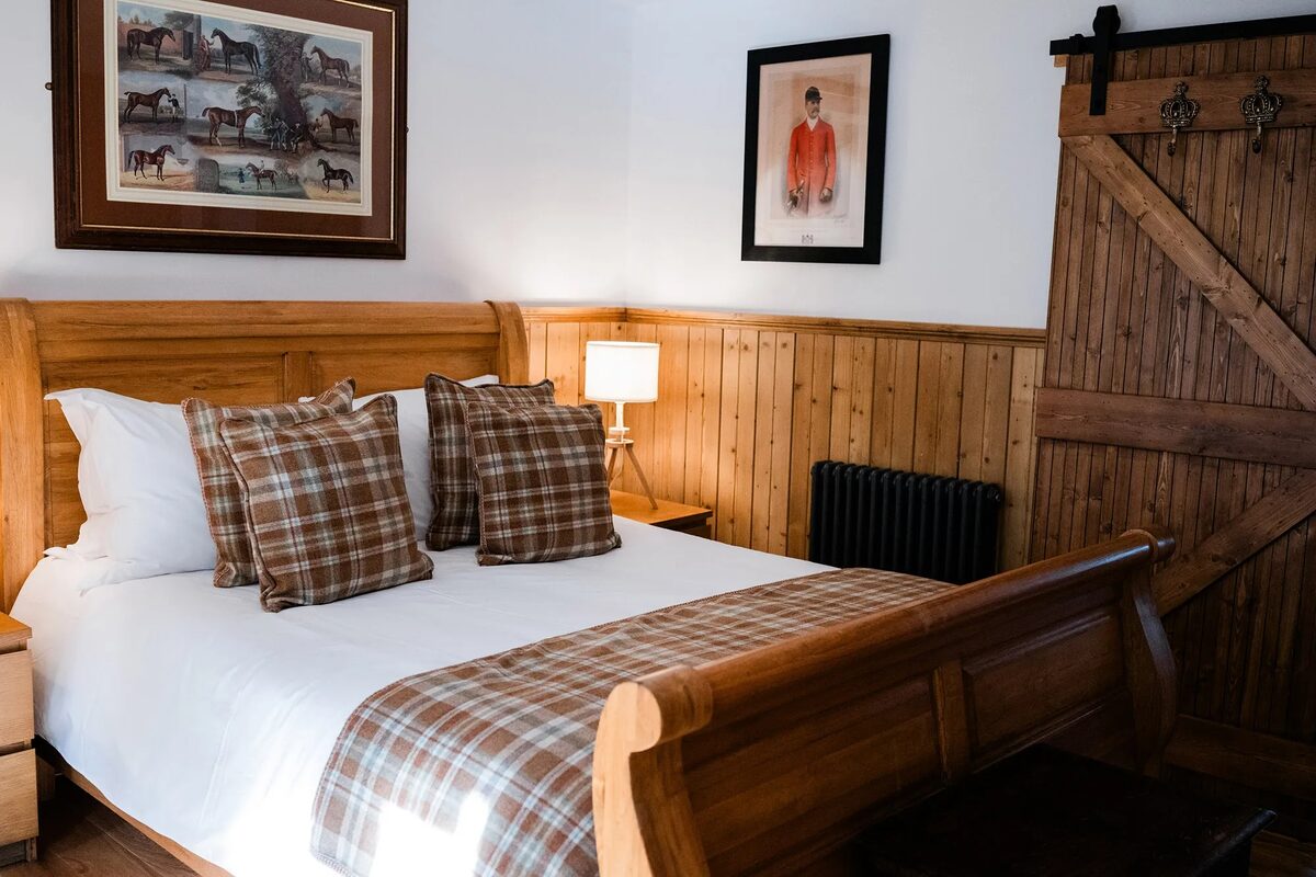 A wooden sleigh bed with tartan cushions sits before panelled walls hung with equestrian prints, a barn door and radiator to the right.