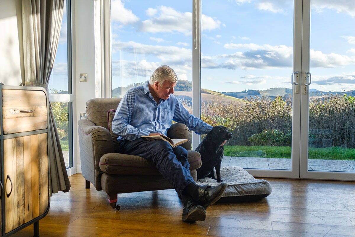 A man reads in an armchair beside a sleeping black dog on a bed, full-height windows looking onto hills behind them.