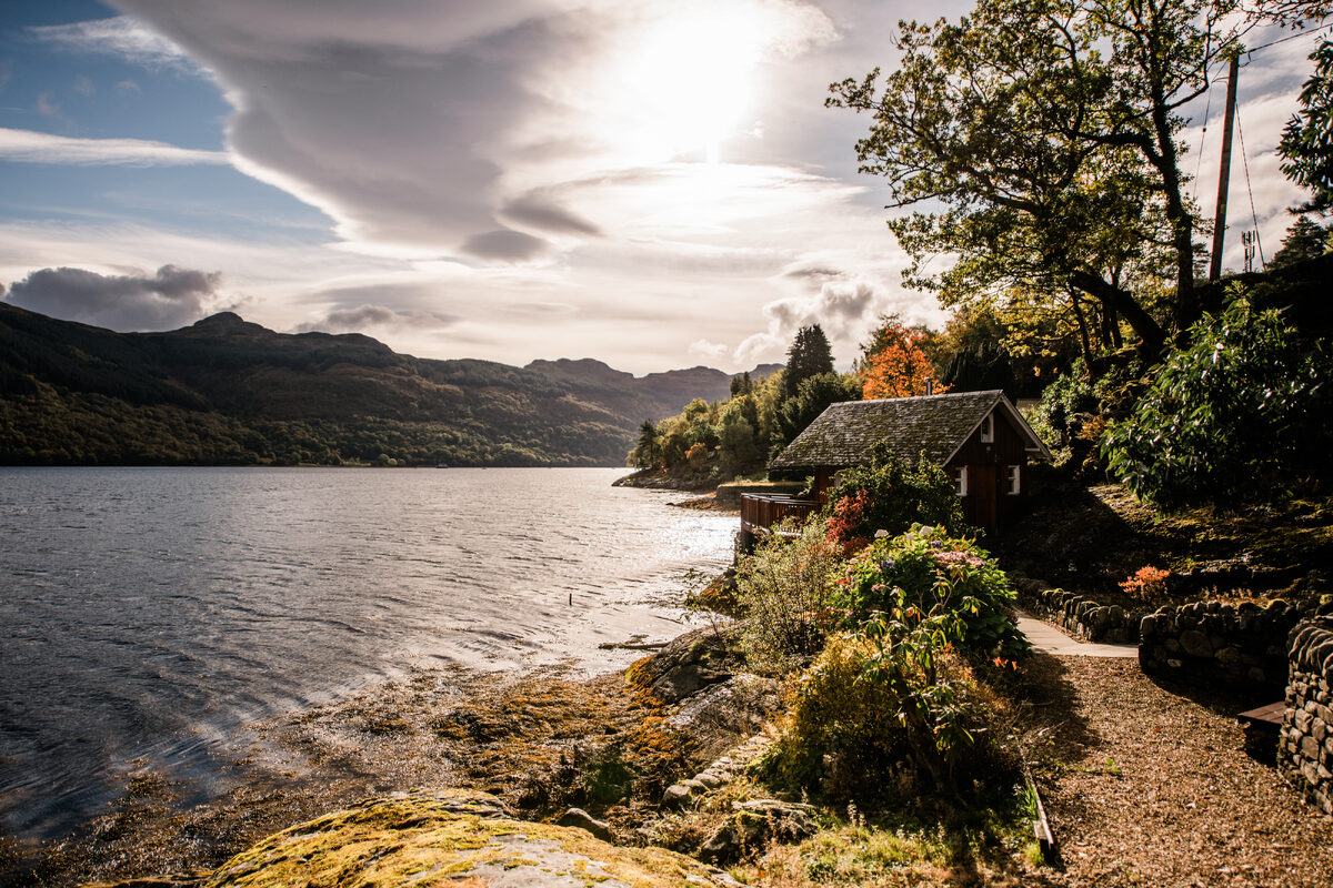 A wood burning stove flue rises from a dark timber cabin on a loch shore, a moored boat visible on the water and hills beyond.