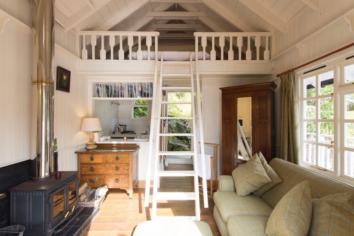 A wooden ladder leads up to a railed mezzanine loft, a wood burning stove, chest of drawers and cream sofa filling the room below.