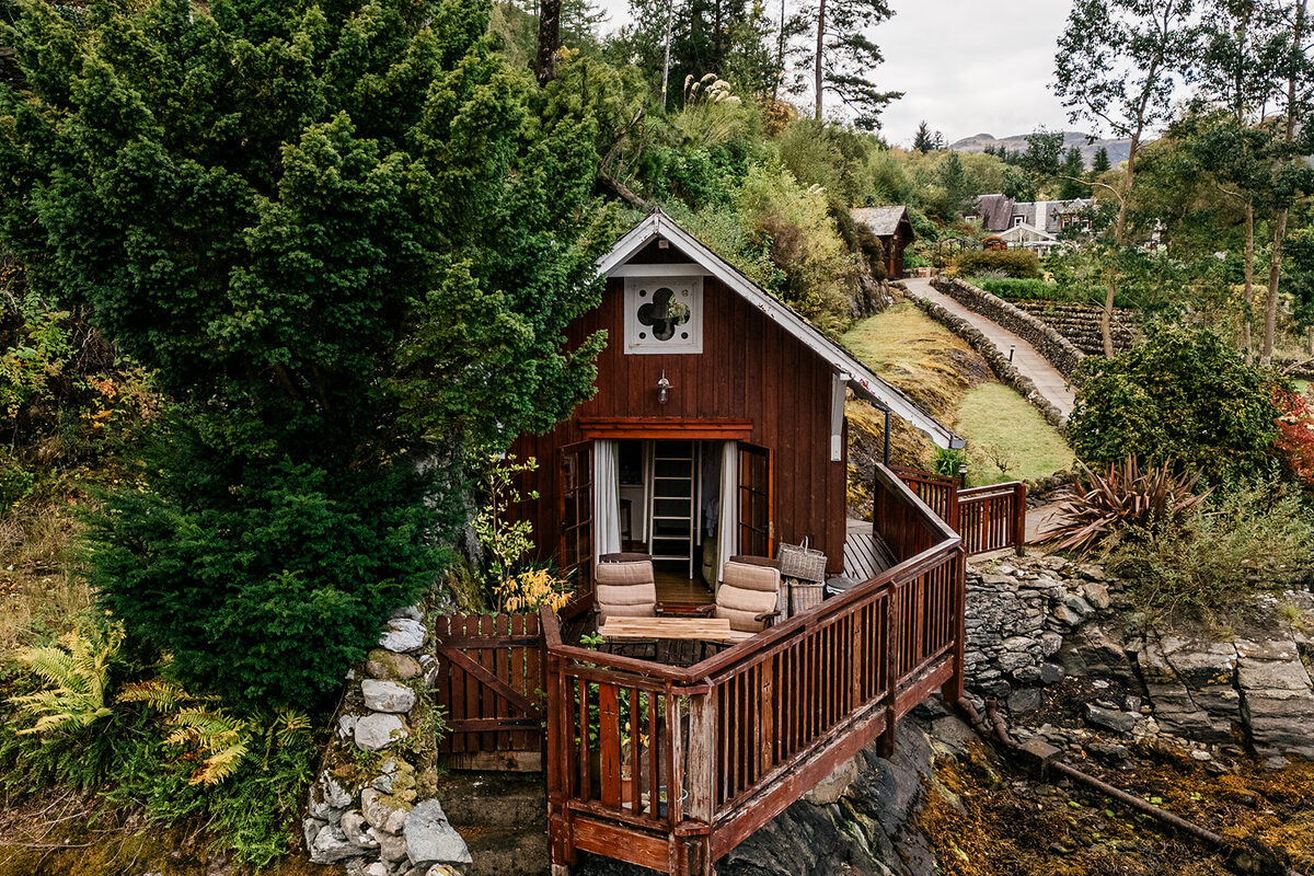 A dark timber cabin with a turf roof and carved gable end sits on a deck overlooking rocky woodland, chairs set outside the glazed door.