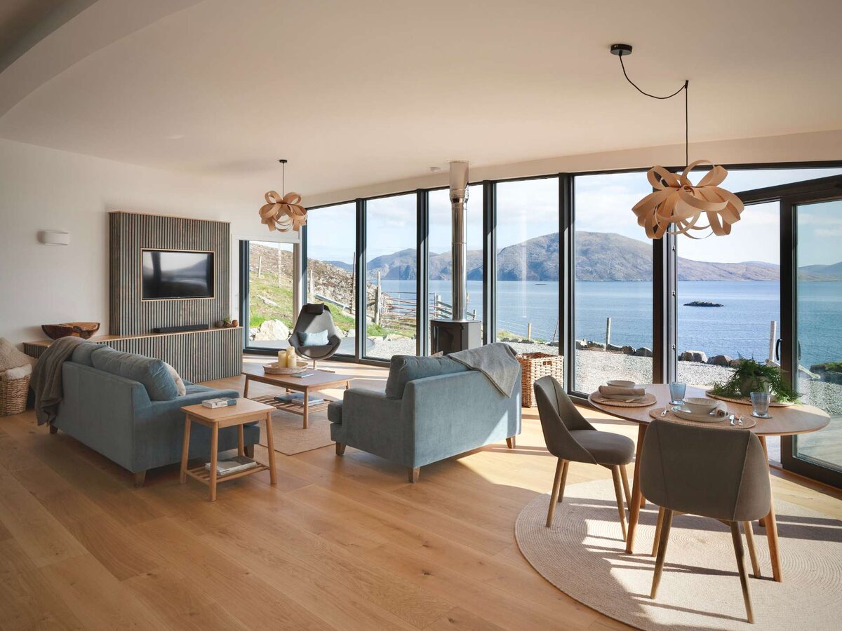 A curved sitting and dining room with grey sofas and a round table looks out through full-height windows onto a sea loch and hills.