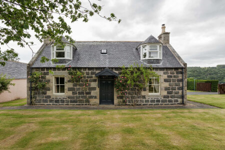 A grey stone cottage with dormer windows and a black front door, climbing roses either side, set behind a lawn.