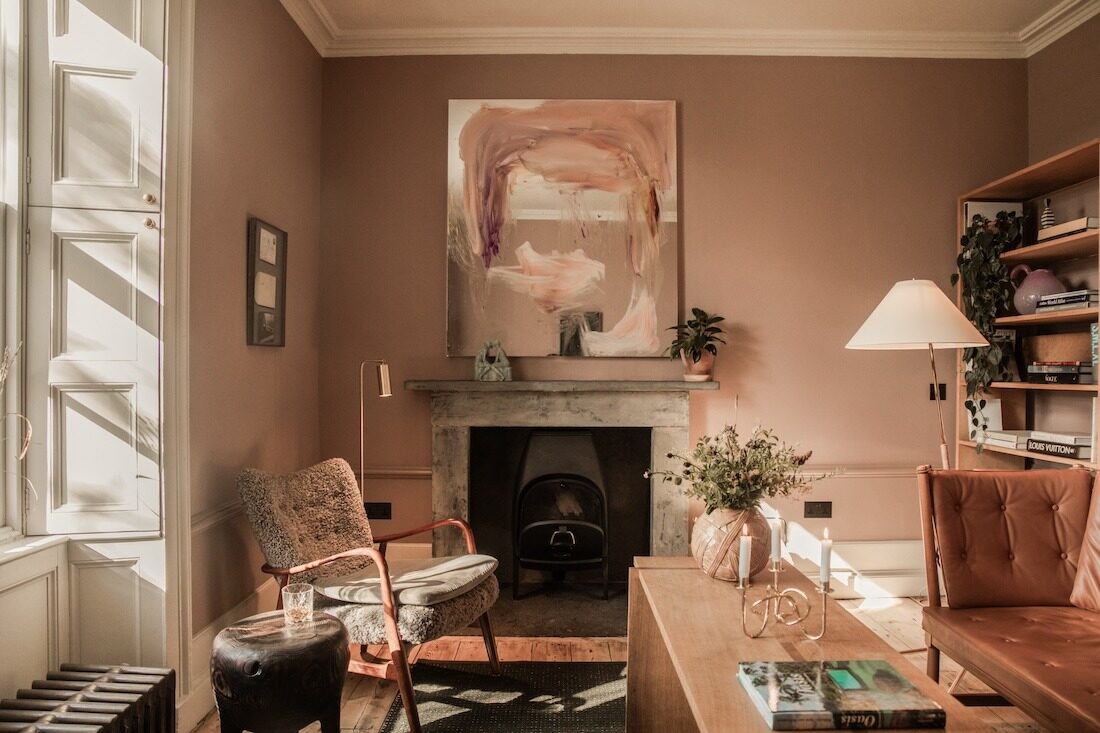A wood burning stove sits in a stone fireplace below a large abstract painting, leather armchairs and a bookshelf completing the room.