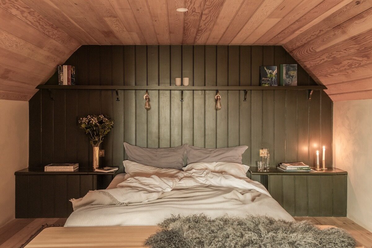 A bed with grey and white bedding stands against a dark green panelled wall beneath a sloped wooden ceiling, lit candles and a small vase of flowers on the bedside shelves.
