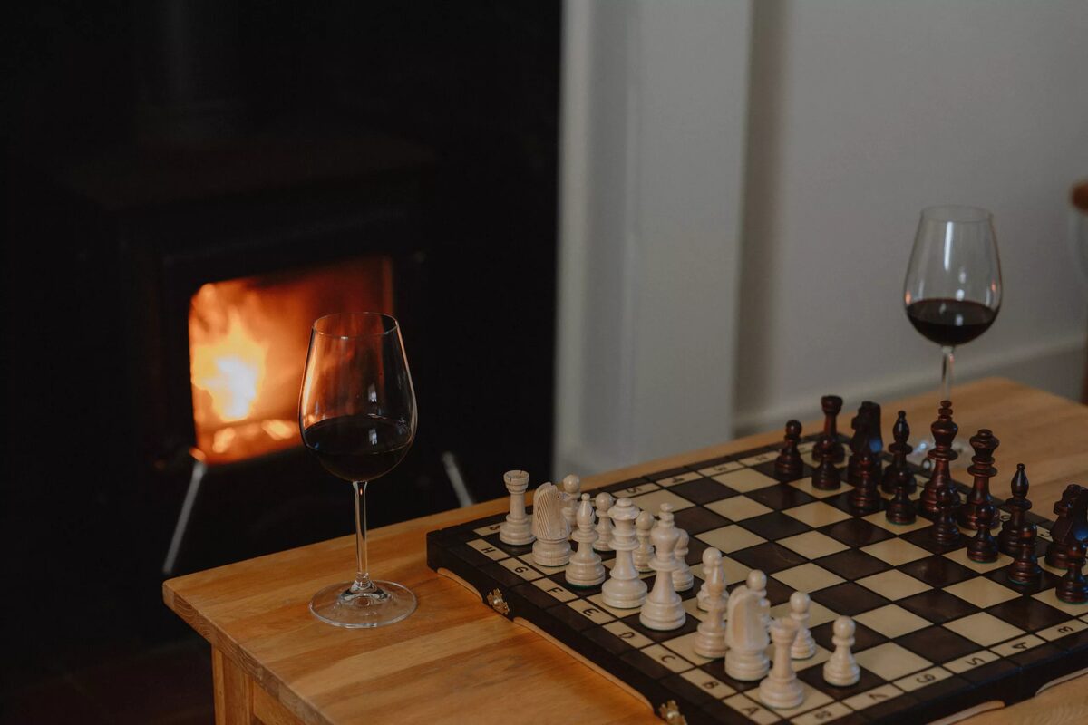 A glass of red wine sits beside a chess set on a low table, a wood burning stove alight in the background.