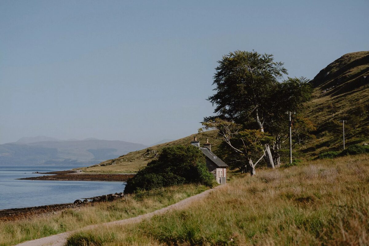 A gravel path leads past a stone cottage towards a sea loch, hills visible across the water under a clear sky.