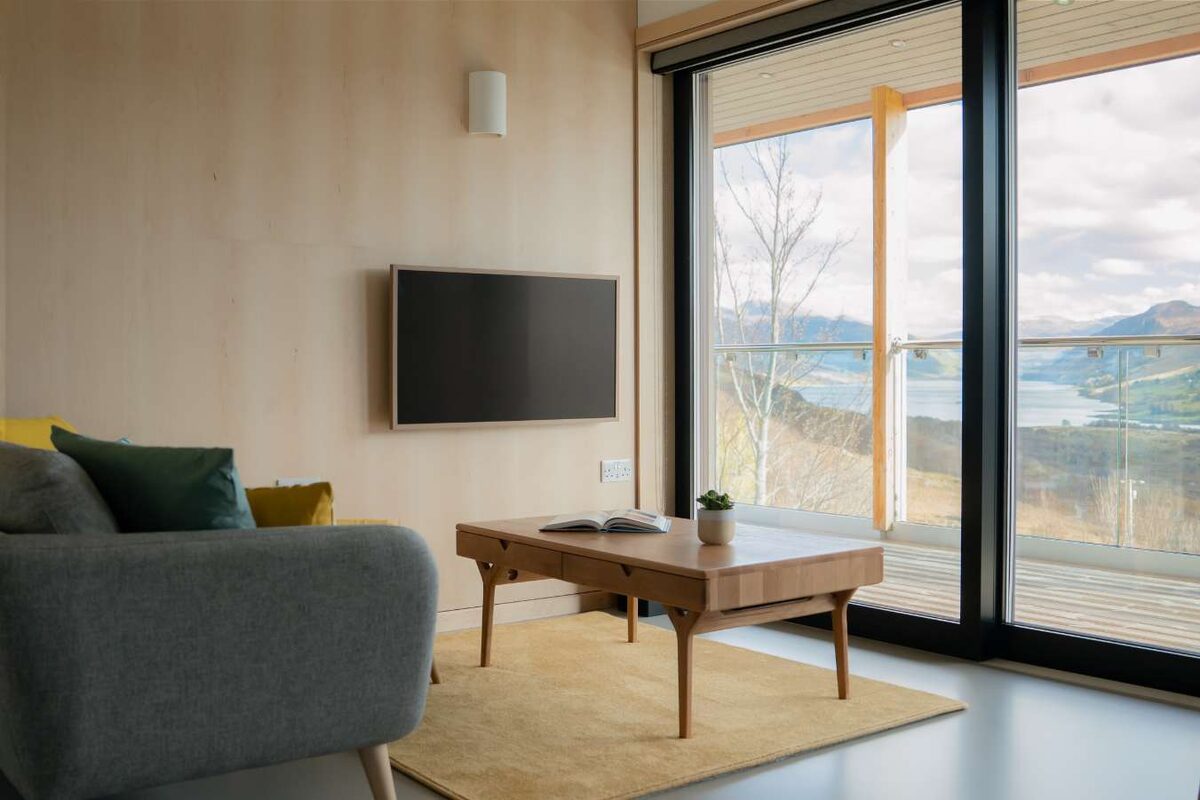 A grey sofa faces a mounted television, full-height glazed doors opening onto a deck with a loch and hills beyond.